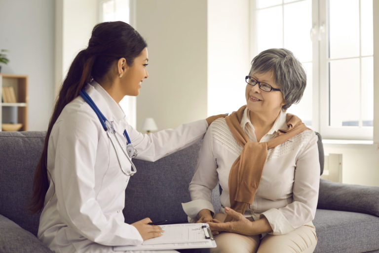 Doctor with clipboard reassuring a smiling senior patient during an in-home visit.