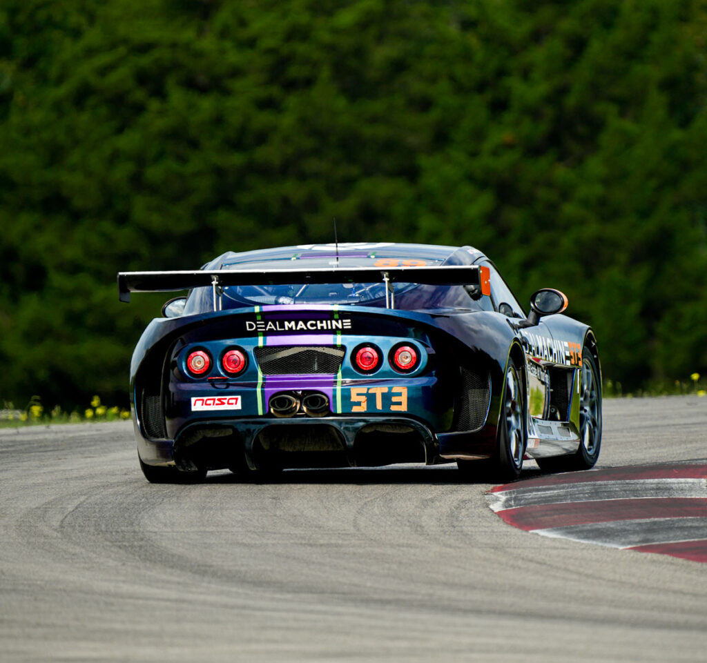 Rear view of the DealMachine-branded racecar cornering on the track with its wrap and spoiler visible.