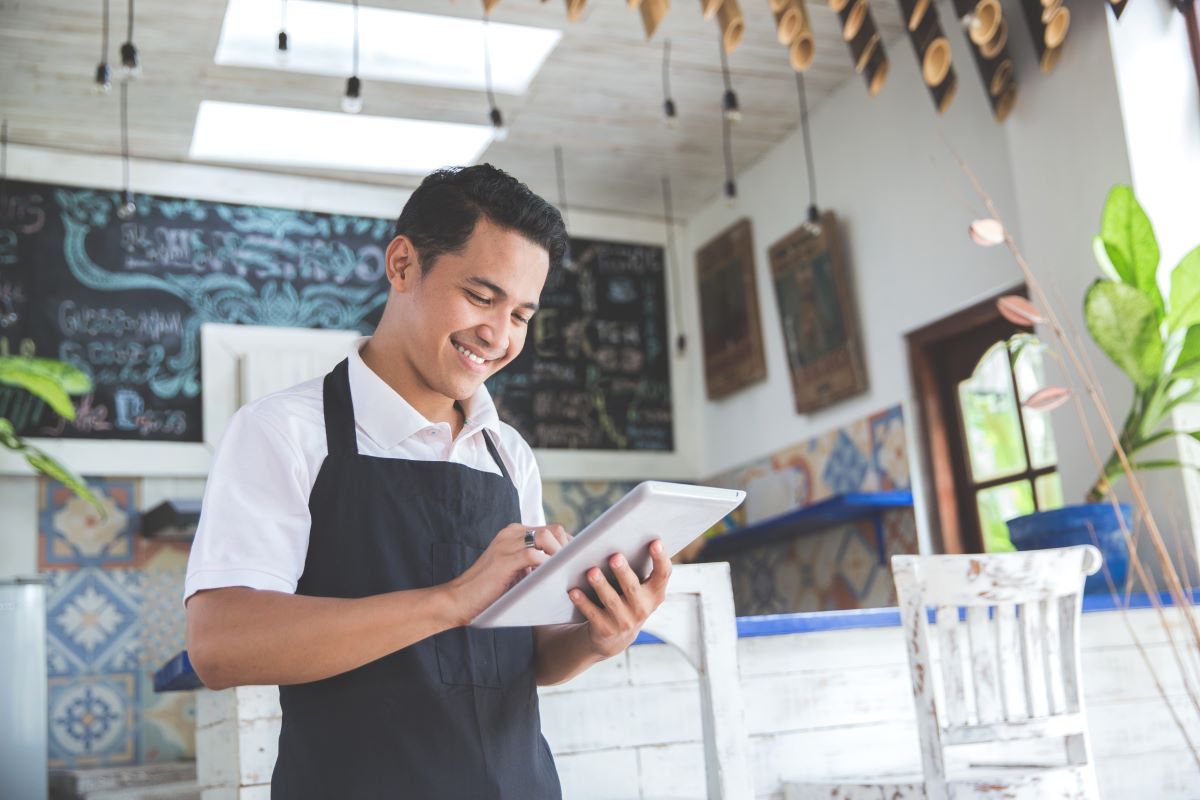 Smiling café employee in an apron using a tablet at the counter.