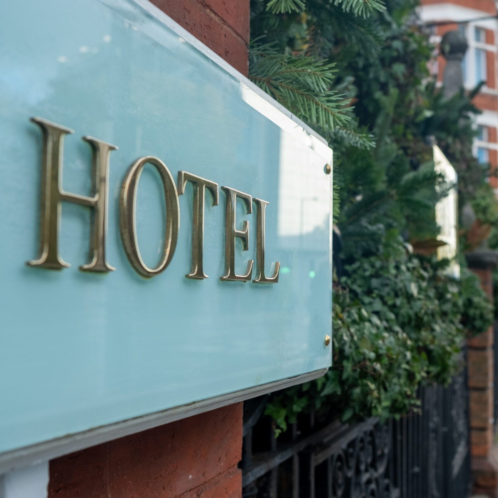 Gold "Hotel" lettering on a teal glass sign against a brick exterior with greenery.