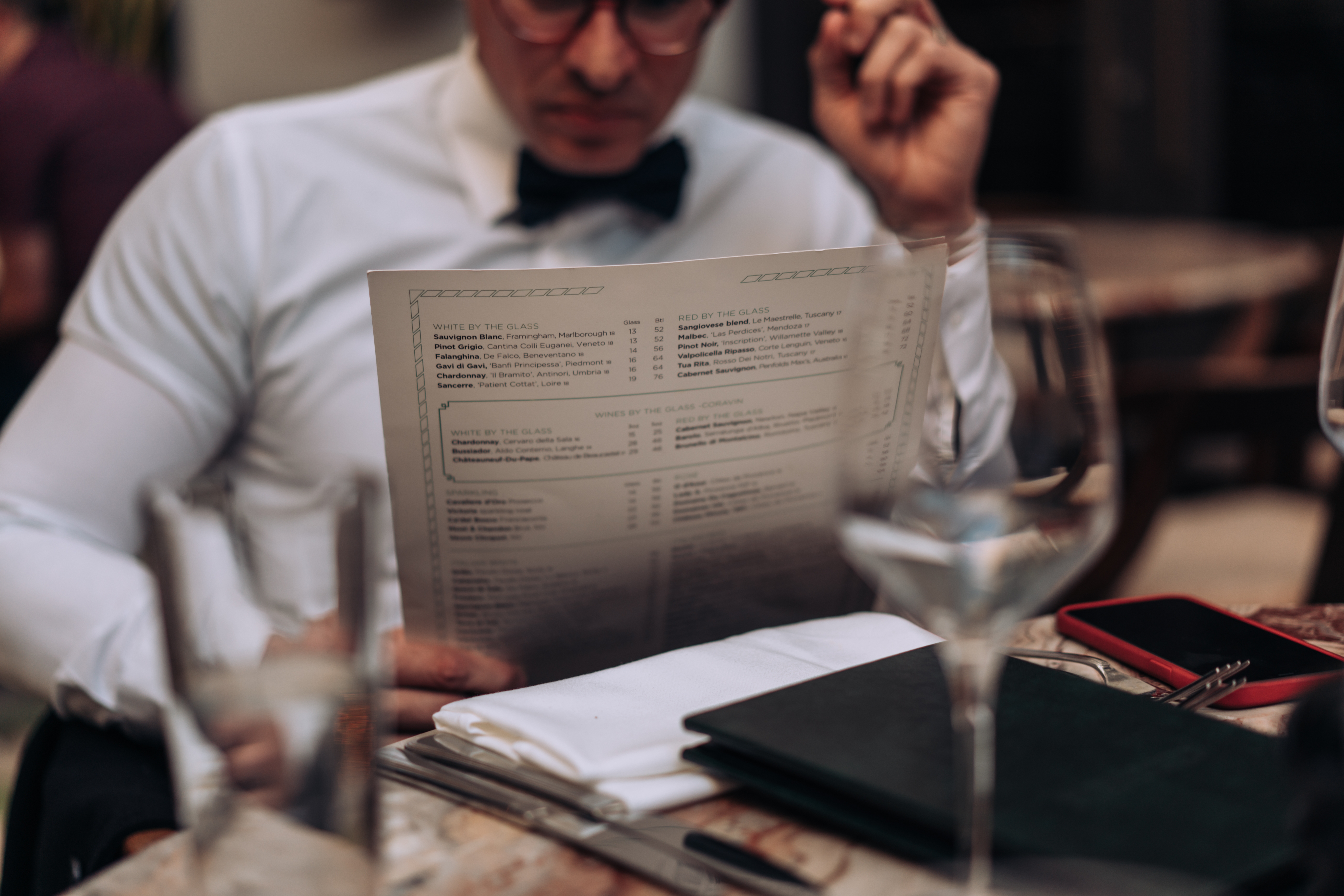 A well-dressed diner reading a printed restaurant menu at a set table.