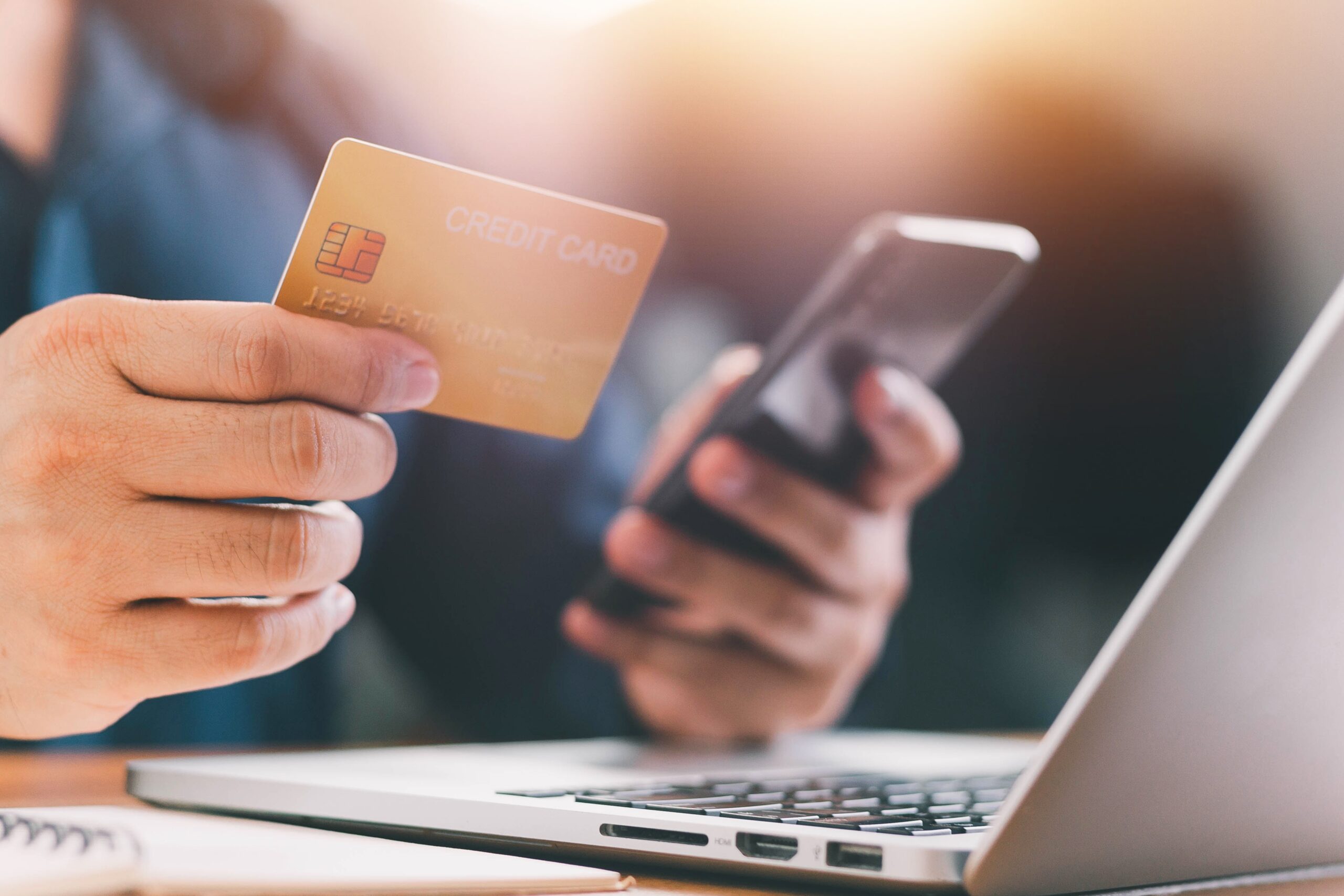 Person holding a credit card while using a phone next to a laptop for an online transaction.