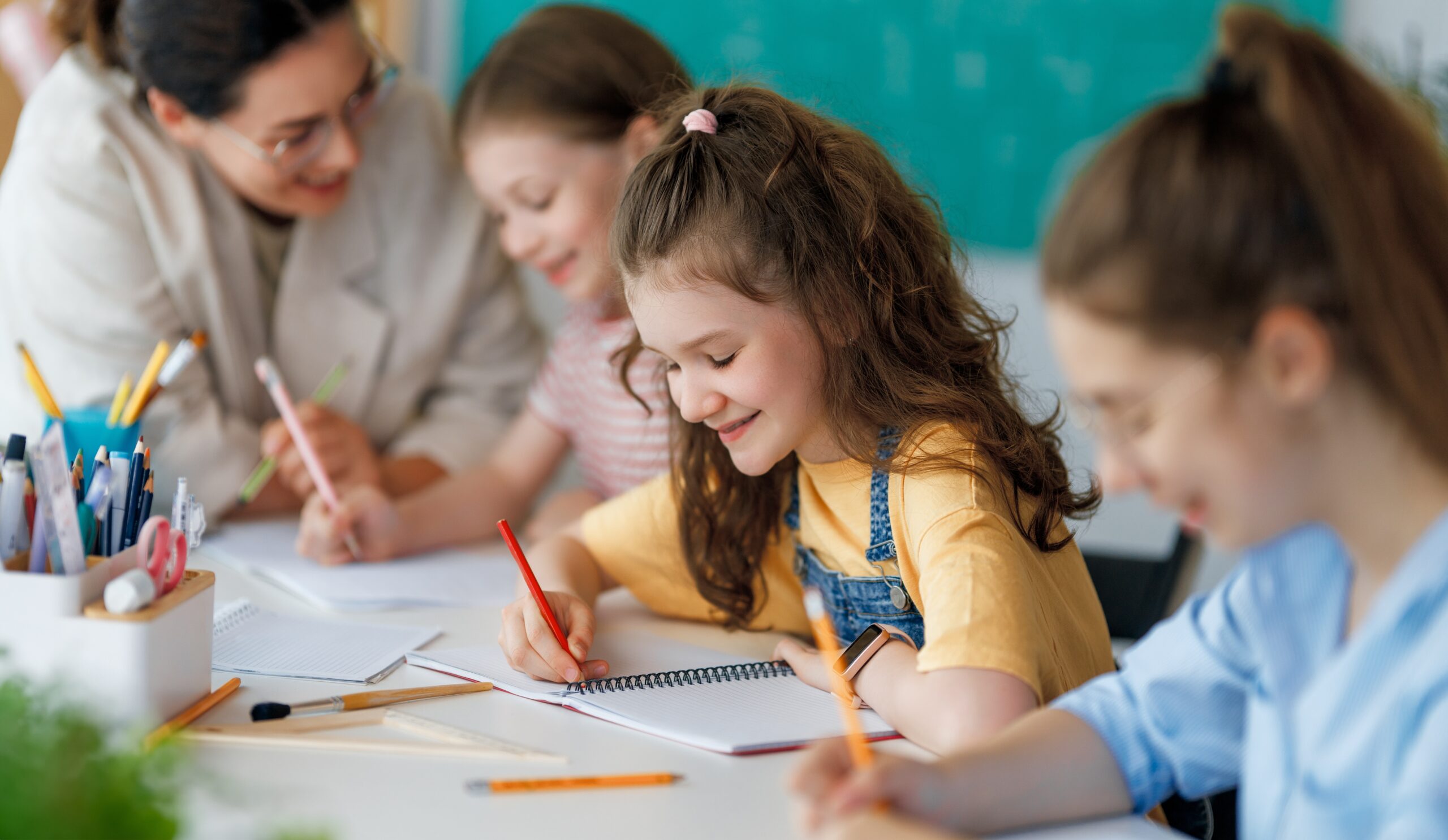 Young students writing at their desks in a classroom with a teacher assisting.