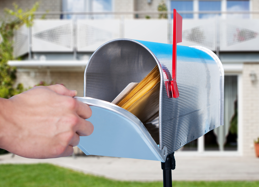 Hand opening a residential mailbox filled with mail outside a home.