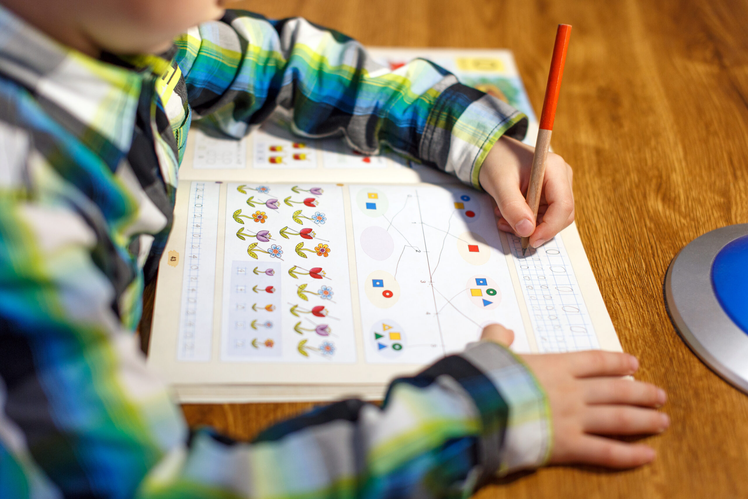 Young child writing in a colorful printed workbook at a wooden table.